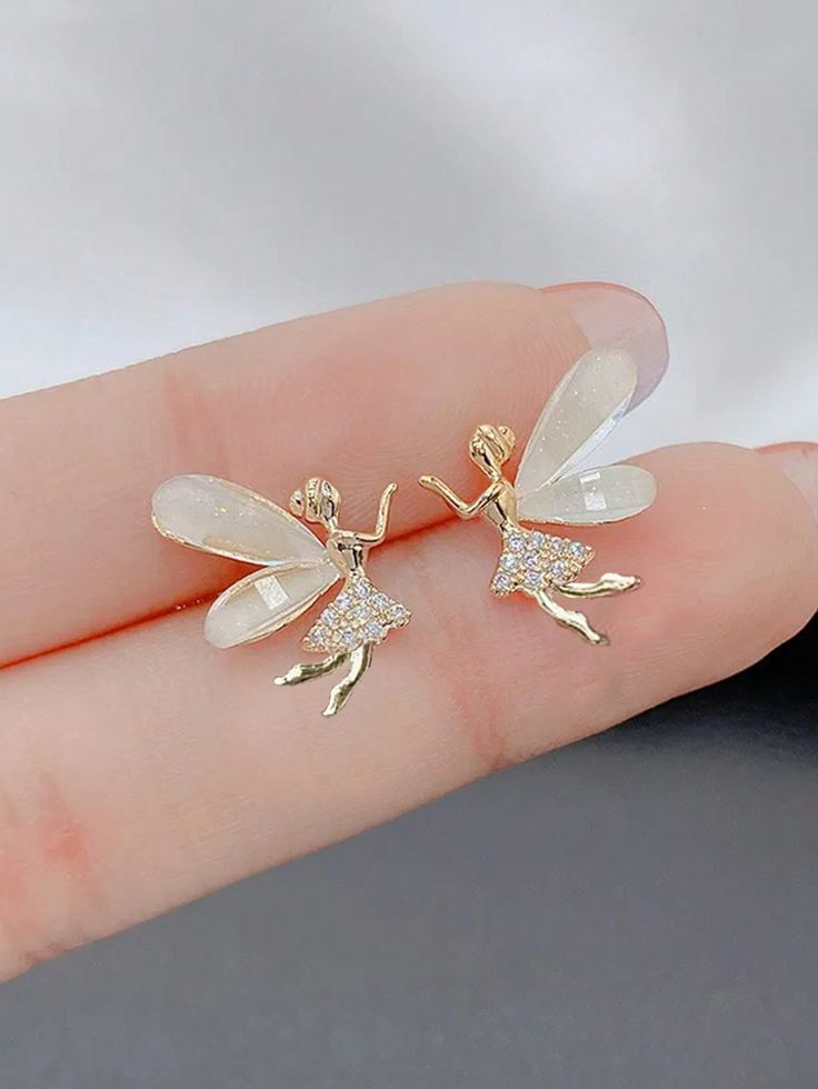 A pair of fairy-shaped stud earrings with gold-toned bodies and translucent white wings, shown resting on a person's fingers against a soft gray background.