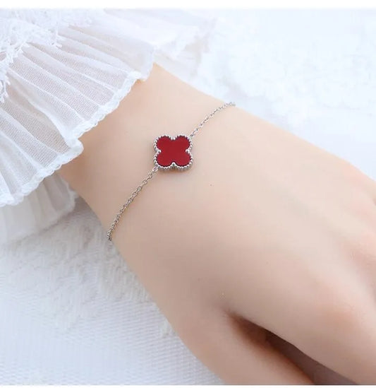 A delicate silver chain bracelet with a red four-leaf clover charm, shown worn on a wrist with a white ruffled sleeve visible.