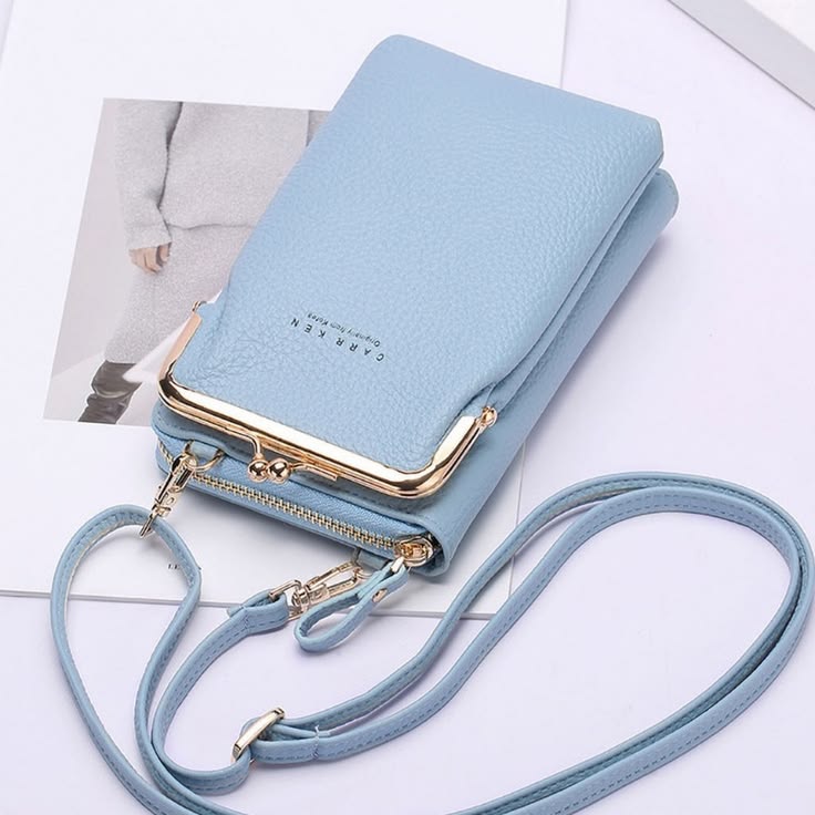 A light blue pebbled leather crossbody wallet with a gold-tone kiss-lock frame and matching strap, displayed on a white surface with a magazine page in the background.